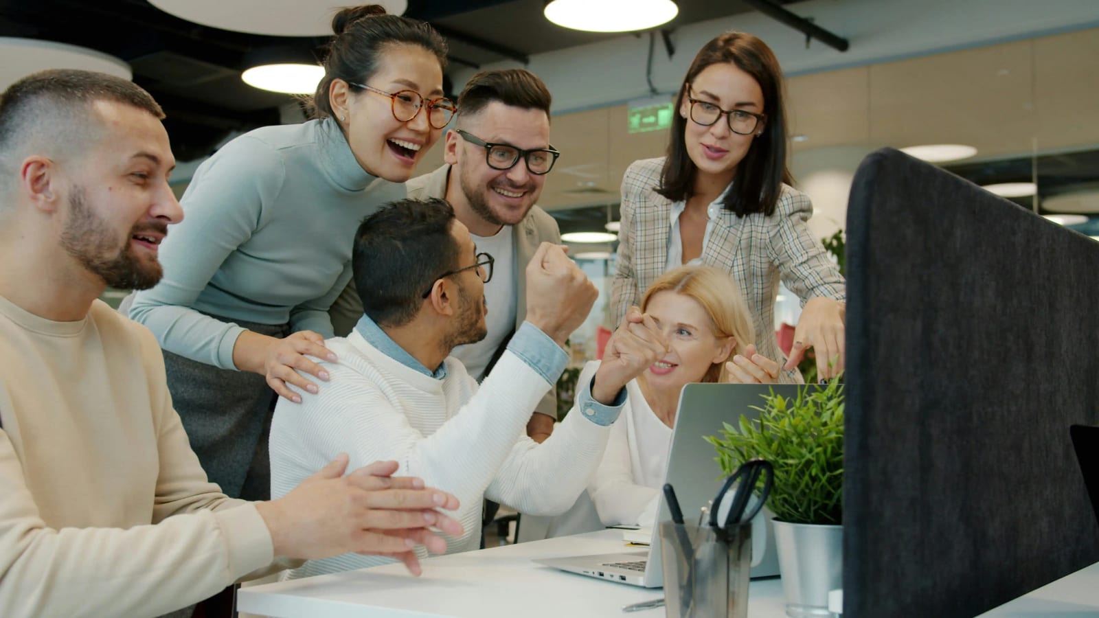A diverse group of colleagues gathered around a laptop in a bright open office, laughing and celebrating together, a candid, energised workplace moment