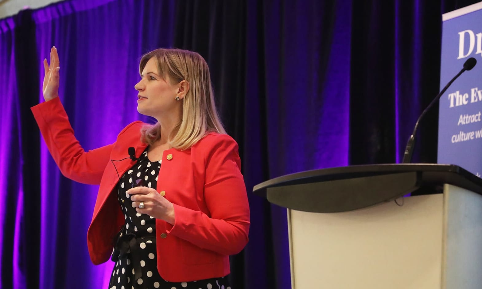 A speaker in a red blazer mid-keynote on a lit conference stage, hand raised as she addresses the room
