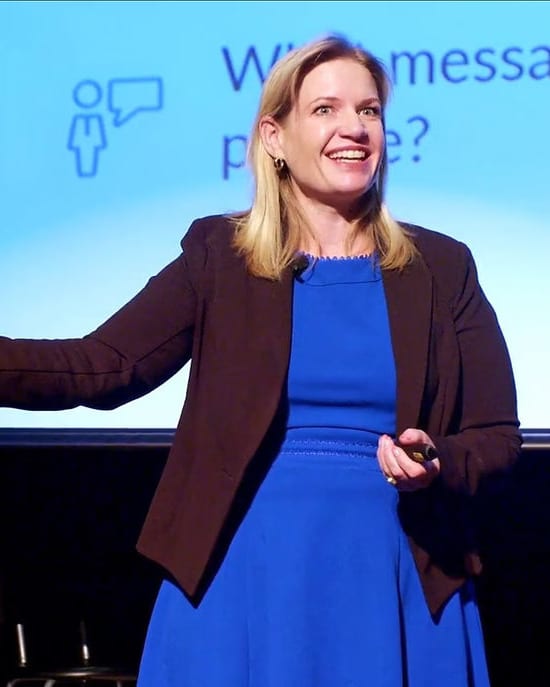 Dr. Laura Hambley presenting on stage mid-gesture, a slide lit up behind her