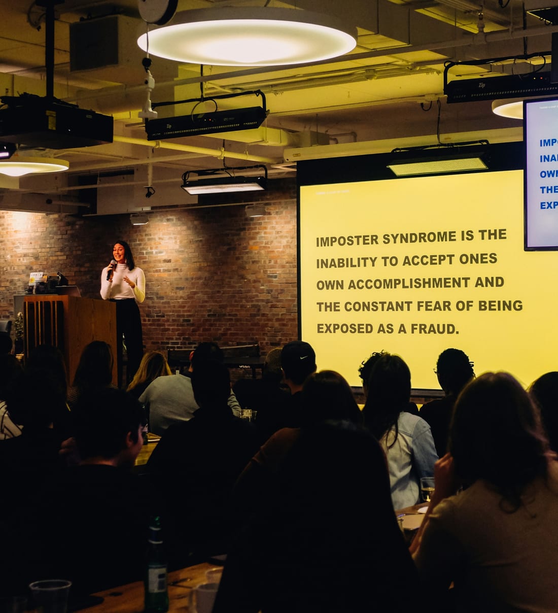 A speaker with a microphone presenting to a seated, engaged audience at a seminar, a bold slide lit up beside her